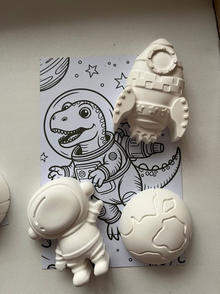 Esculturas de foguete, astronauta e planeta em gesso branco sobre um desenho de dinossauro astronauta